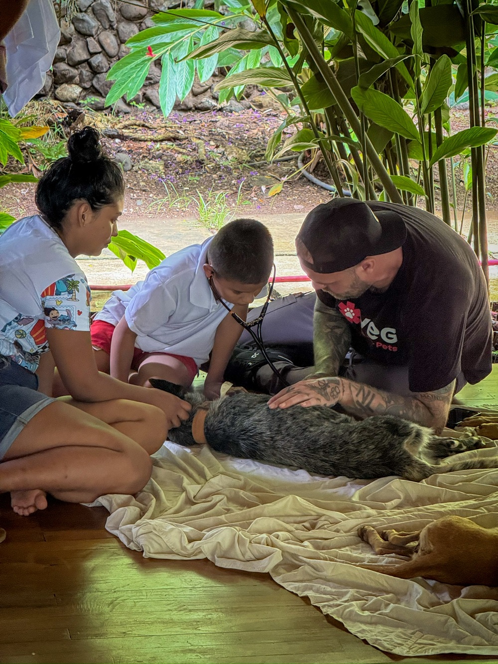 A young boy listens to a cat's heartbeat with a stethoscope, guided by a PRCP volunteer and a local mother