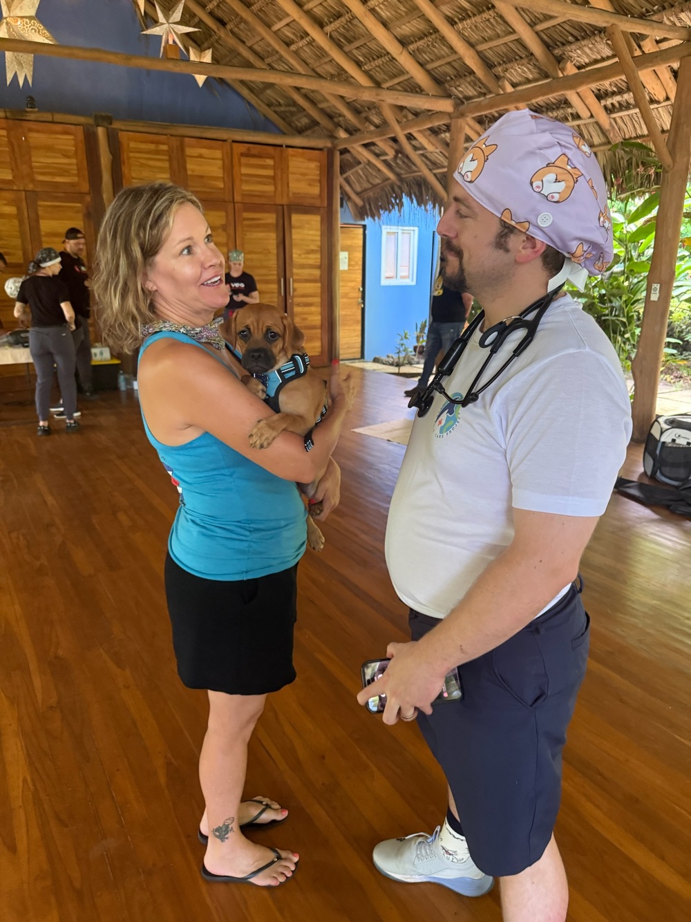 A smiling woman holds her puppy while chatting with a PRCP veterinarian inside the open-air clinic pavilion