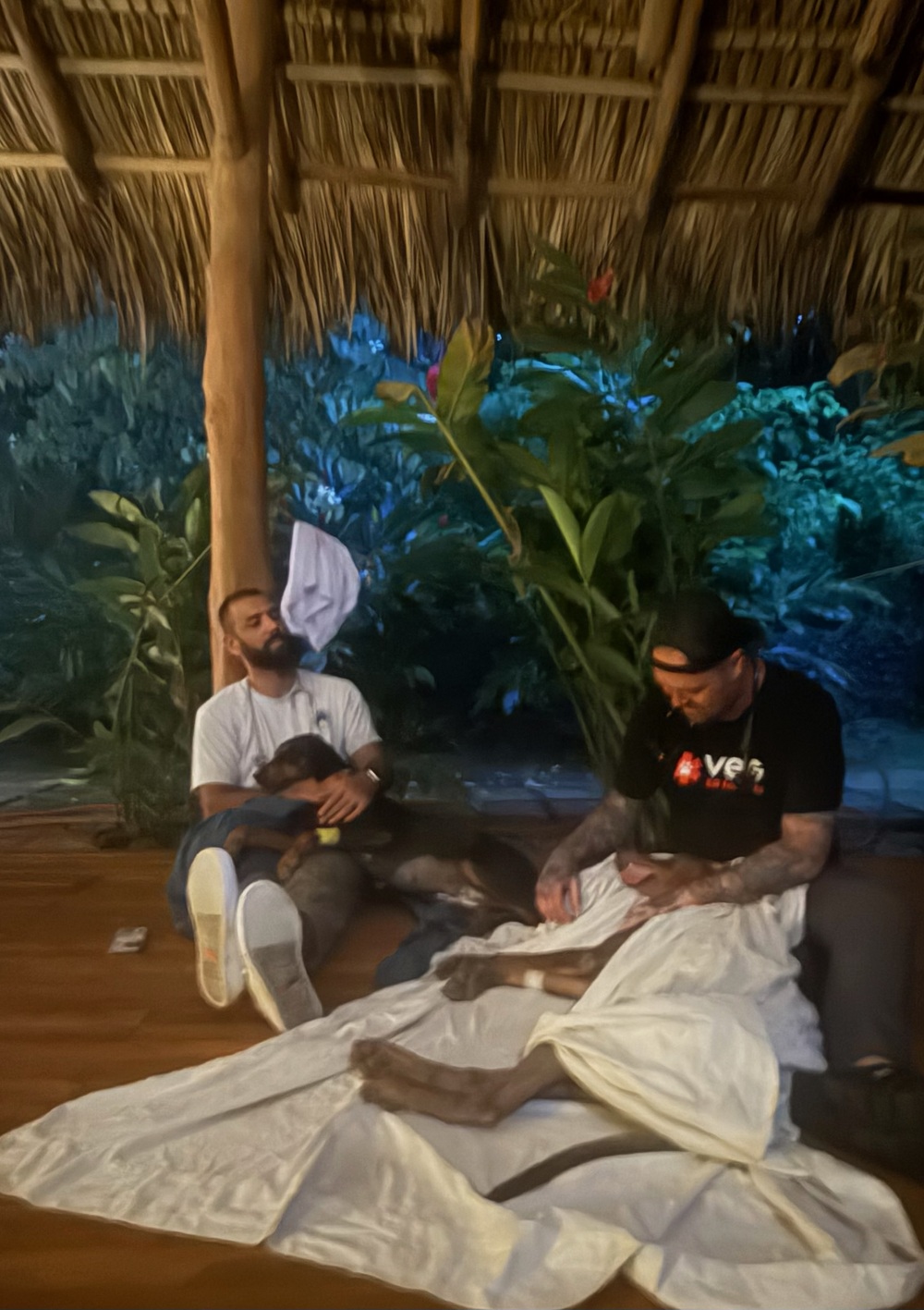 Two PRCP volunteers sit on the floor under an open palapa at night, keeping recovering dogs warm and calm