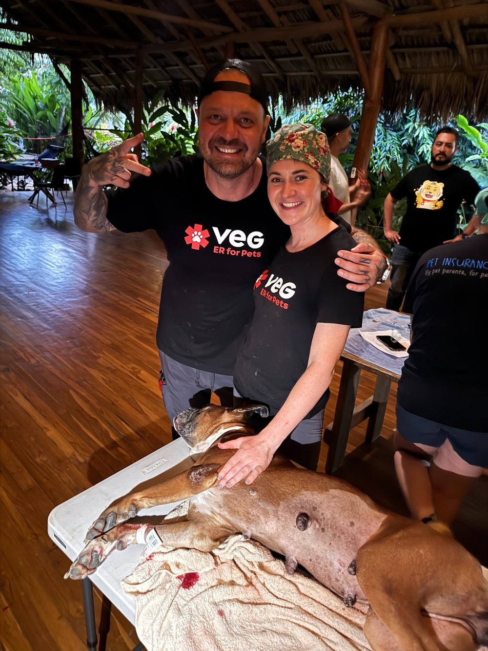 Two VEG volunteers smile together with a dog resting on the exam table after a successful procedure
